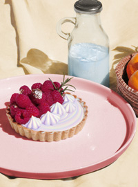 A digital image depicting a large pink plate with a raspberry and pastel rainbow colored cream tart. Behind the plate is a clear jug of blue milk, and a corner of a basket filled with oranges. The background is a yellow cloth.